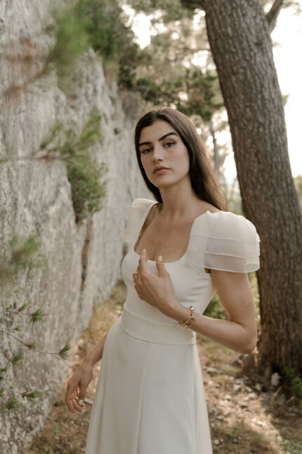 LA ROBE AQUA AURA est une robe de mariée moderne et couture en crêpe de soie. Sa silhouette est longiligne, son buste décolleté carré est habillé de manches courtes gonflantes et sa taille soulignée d’une ceinture finit sur une traine légère en organza, pour une mariée poétique. LA ROBE AQUA AURA