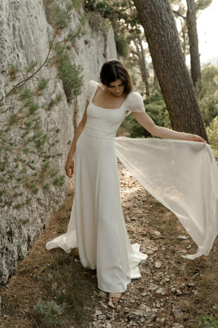 LA ROBE AQUA AURA est une robe de mariée moderne et couture en crêpe de soie. Sa silhouette est longiligne, son buste décolleté carré est habillé de manches courtes gonflantes et sa taille soulignée d’une ceinture finit sur une traine légère en organza, pour une mariée poétique. LA ROBE AQUA AURA