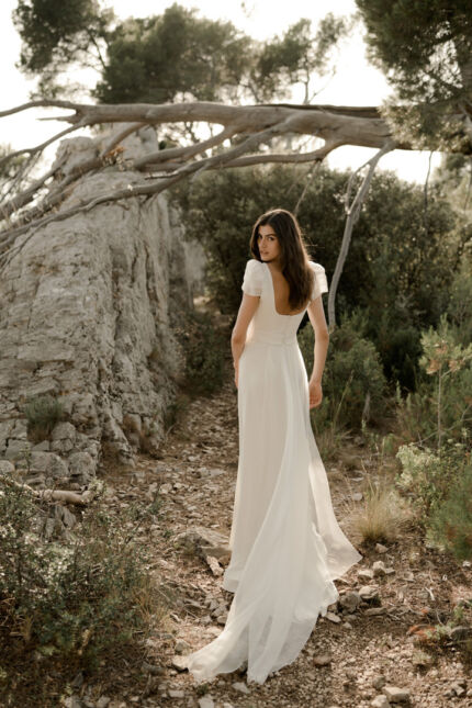 LA ROBE AQUA AURA est une robe de mariée moderne et couture en crêpe de soie. Sa silhouette est longiligne, son buste décolleté carré est habillé de manches courtes gonflantes et sa taille soulignée d’une ceinture finit sur une traine légère en organza, pour une mariée poétique. LA ROBE AQUA AURA