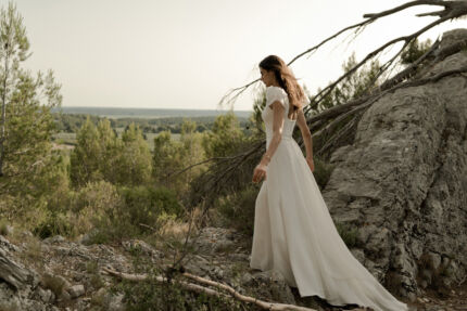 LA ROBE AQUA AURA est une robe de mariée moderne et couture en crêpe de soie. Sa silhouette est longiligne, son buste décolleté carré est habillé de manches courtes gonflantes et sa taille soulignée d’une ceinture finit sur une traine légère en organza, pour une mariée poétique. LA ROBE AQUA AURA