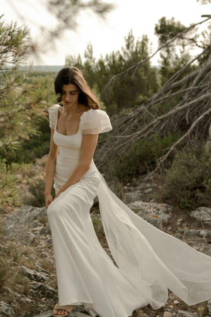 LA ROBE AQUA AURA est une robe de mariée moderne et couture en crêpe de soie. Sa silhouette est longiligne, son buste décolleté carré est habillé de manches courtes gonflantes et sa taille soulignée d’une ceinture finit sur une traine légère en organza, pour une mariée poétique. LA ROBE AQUA AURA