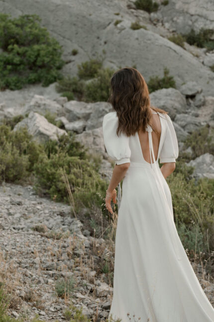 LA ROBE AMBRE est une robe de mariée moderne chic et couture toute en crêpe de soie. Des manches gonflantes à plis dessinent les épaules, son grand décolleté sensuel et sa fente conviendront à une mariée amazone des temps modernes. LA ROBE AMBRE