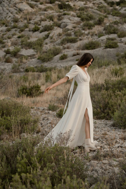 LA ROBE AMBRE est une robe de mariée moderne chic et couture toute en crêpe de soie. Des manches gonflantes à plis dessinent les épaules, son grand décolleté sensuel et sa fente conviendront à une mariée amazone des temps modernes. LA ROBE AMBRE