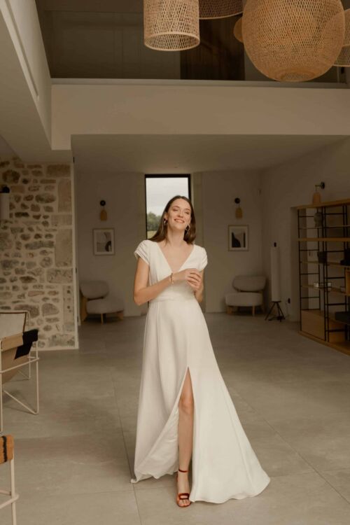 NIMIO est une robe de mariée moderne et chic, en crêpe de soie ornée d'une ceinture et de petites manches drapées.