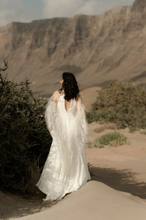 PARADISE est une robe de mariée chic en crêpe de soie. Son décolleté « emmanchures américaines » et son dos habillé de bretelles en font une robe de mariée minimaliste élégante. La jupe est incrustée de mousseline de soie pour faire une robe de mariée fluide. Bijoux @anthemis.paris . « Plumes » est une étole de mariée en broderie de plumes.
