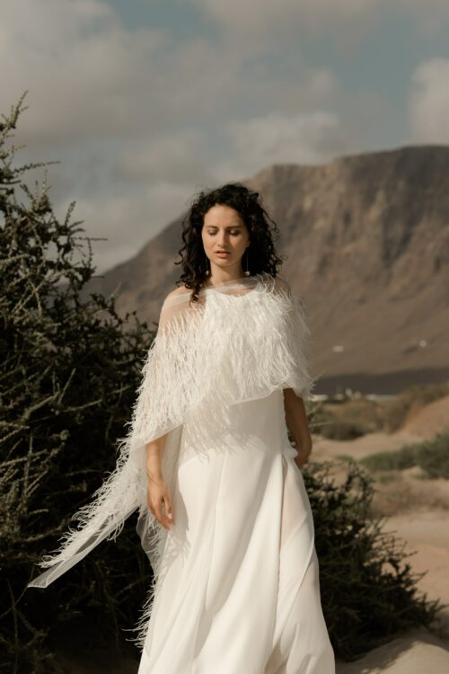 PARADISE est une robe de mariée chic en crêpe de soie. Son décolleté « emmanchures américaines » et son dos habillé de bretelles en font une robe de mariée minimaliste élégante. La jupe est incrustée de mousseline de soie pour faire une robe de mariée fluide. Bijoux @anthemis.paris . « Plumes » est une étole de mariée en broderie de plumes.