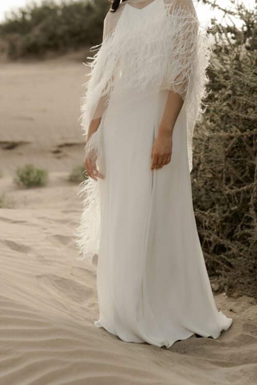 PARADISE est une robe de mariée chic en crêpe de soie. Son décolleté « emmanchures américaines » et son dos habillé de bretelles en font une robe de mariée minimaliste élégante. La jupe est incrustée de mousseline de soie pour faire une robe de mariée fluide. Bijoux @anthemis.paris . « Plumes » est une étole de mariée en broderie de plumes.